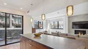 A modern kitchen with a large island, wooden cabinets, pendant lighting, and a stone fireplace. - The Lion at Vail W302 (Vail)