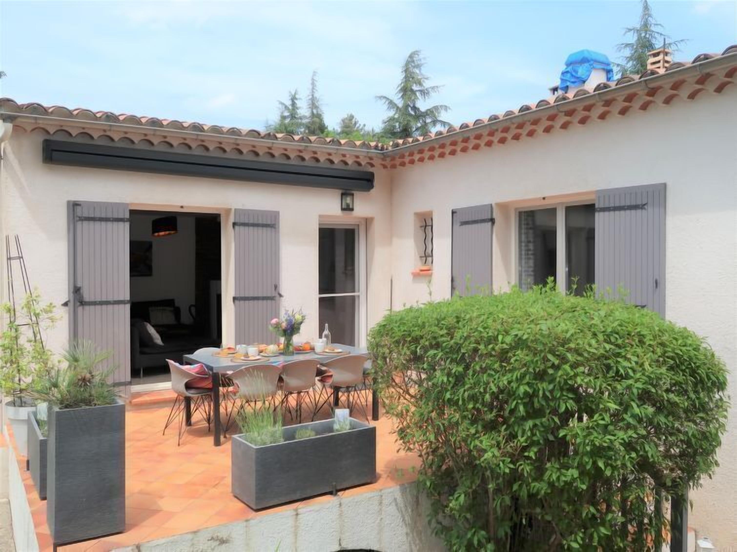 A patio with a table and chairs, a small garden, and a building with a tiled roof and shutters.