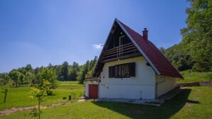 Uma pequena casa térrea com telhado vermelho e paredes brancas, cercada por um campo gramado e árvores.