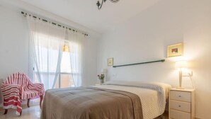 A bedroom with a bed, a nightstand, a chair, and a window with sheer curtains. - Bosc de la Montserrada by Interhome (La Pineda)