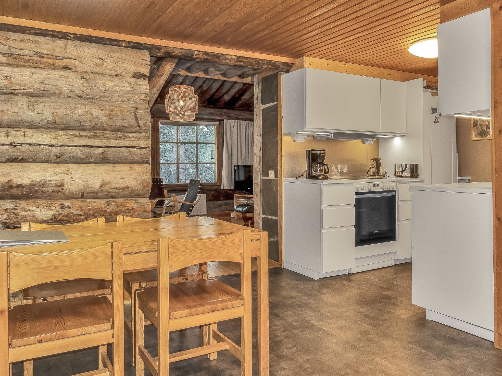 A rustic cabin interior with a wooden dining table, chairs, a kitchen with white cabinets, and a large window.