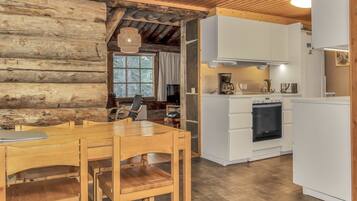 A rustic cabin interior with a wooden dining table, chairs, a kitchen with white cabinets, and a large window.