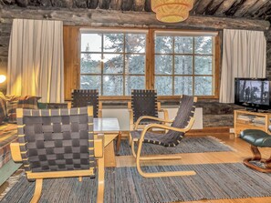 Ein gemütliches Blockhaus-Interieur mit einem Holztisch, zwei Stühlen, einem Fernseher und einem großen Fenster mit Blick auf Bäume.