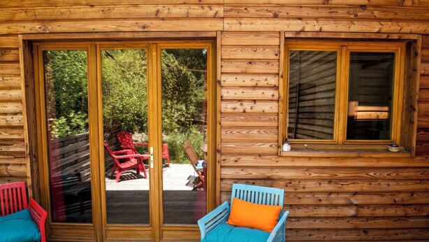 A wooden cabin with a sliding glass door, a deck with red and blue chairs, and a window. - Chalet Jardin Zaza (saint remèze)