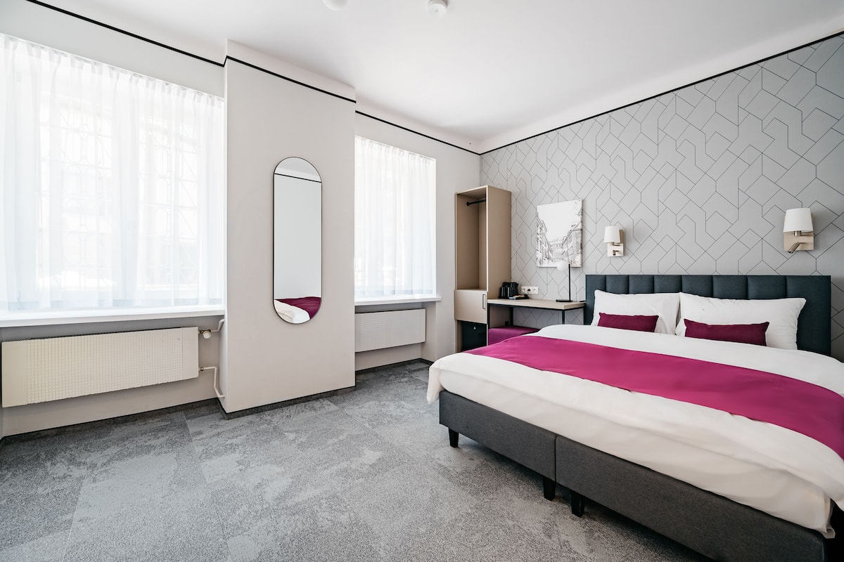 Una habitación de hotel moderna con una cama grande, una mesita de noche, un espejo y una ventana con cortinas translúcidas.