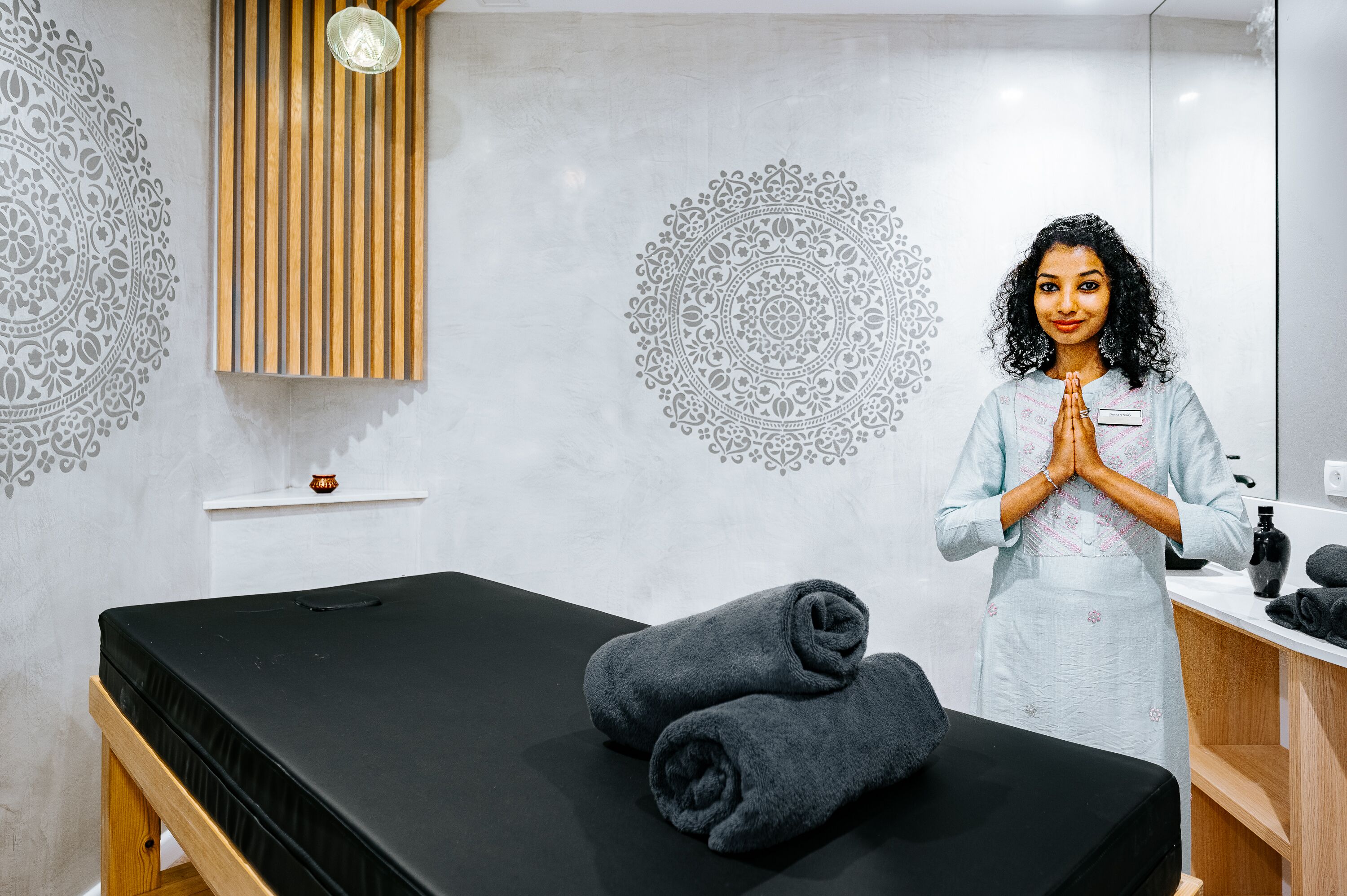 A massage therapy room with a black massage table, two rolled-up towels, and a wooden panel with vertical slats.