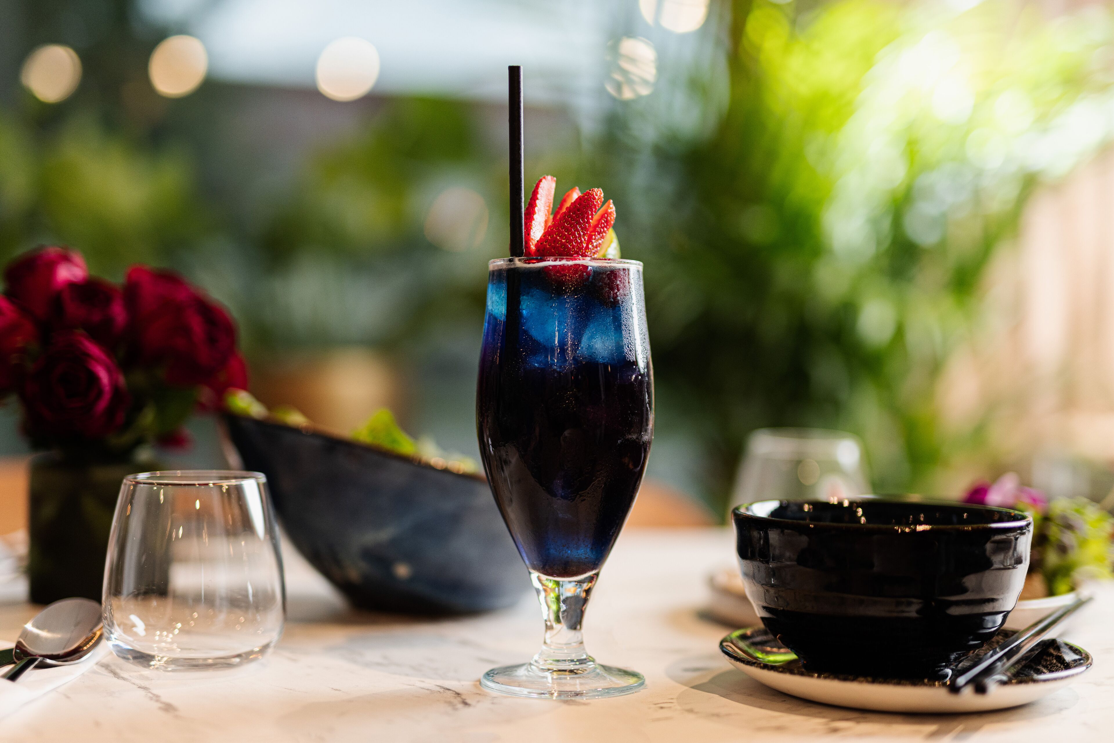Un vaso con una bebida oscura y una fresa como adorno, un vaso transparente y un cuenco negro sobre una mesa.