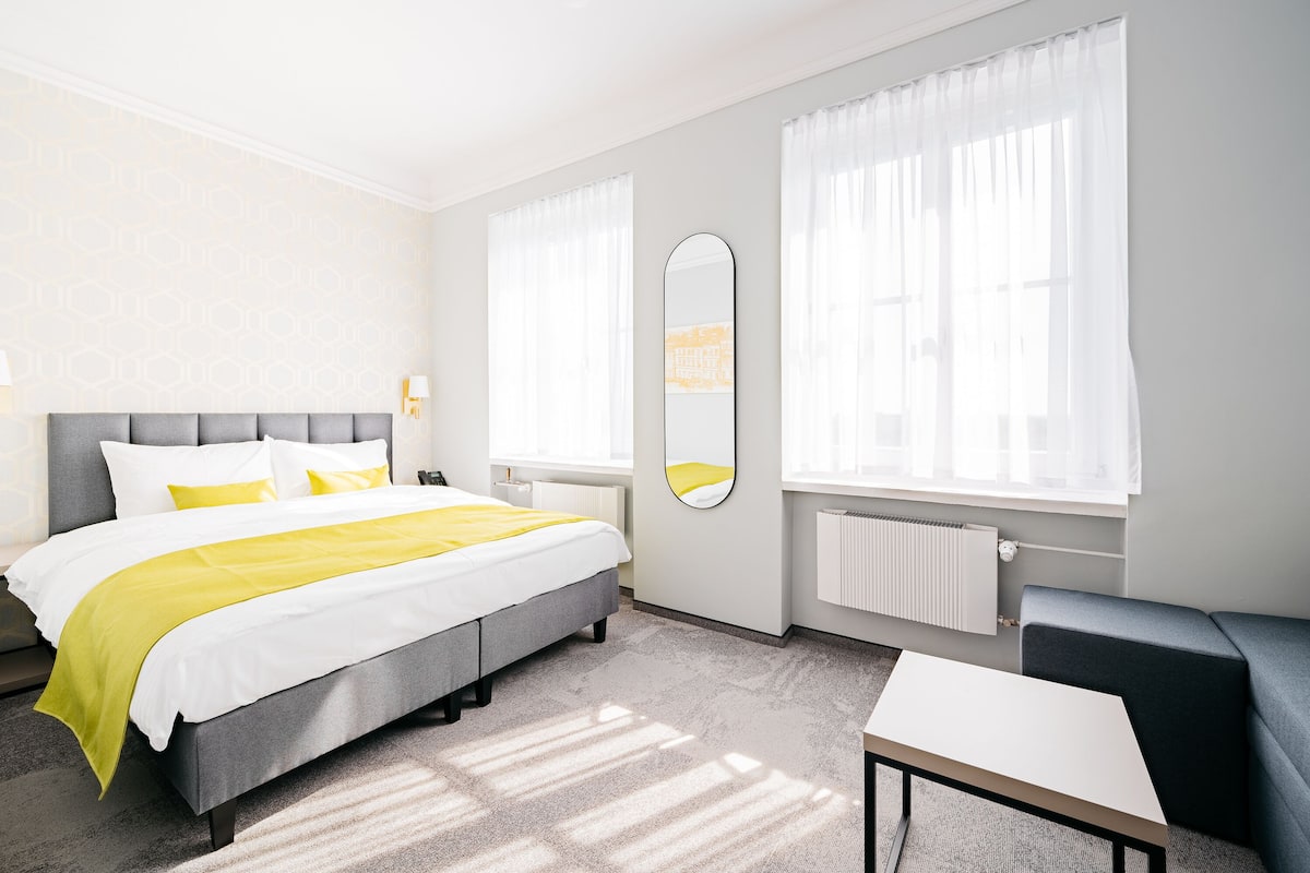Una habitación de hotel moderna con una cama grande, una mesita de noche, una silla y un ventanal con cortinas.