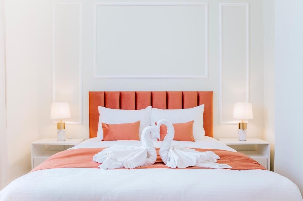 Cama bem arrumada com cabeceira, duas luminárias de apoio e arranjo de toalha em formato de cisne.