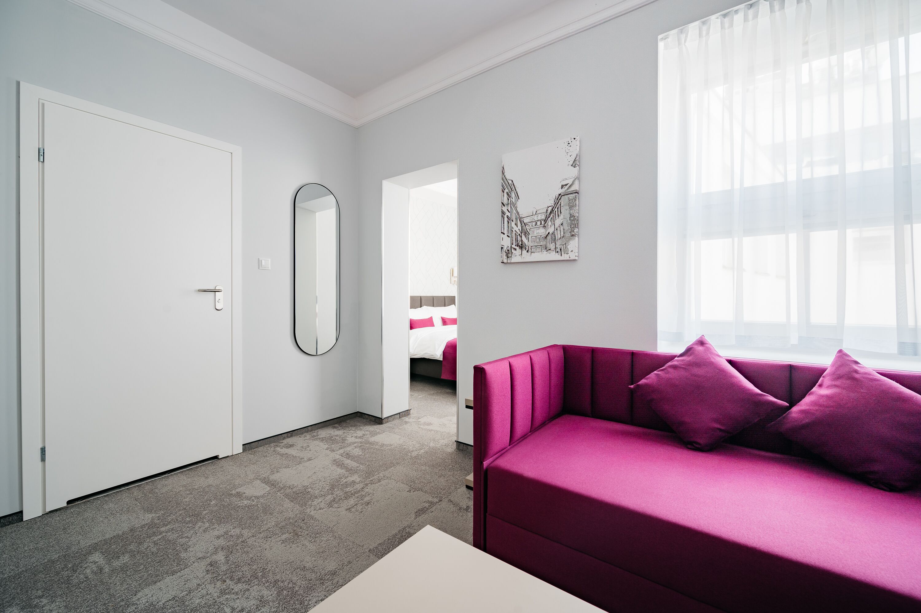 Une pièce moderne avec un canapé rose, un grand miroir et une œuvre d’art représentant un paysage urbain accrochée au mur.