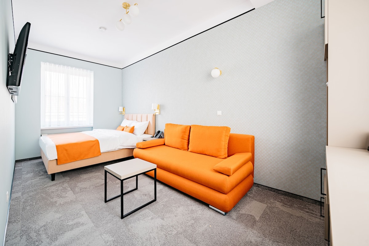 Una habitación de hotel moderna con cama, sofá naranja y una mesita.