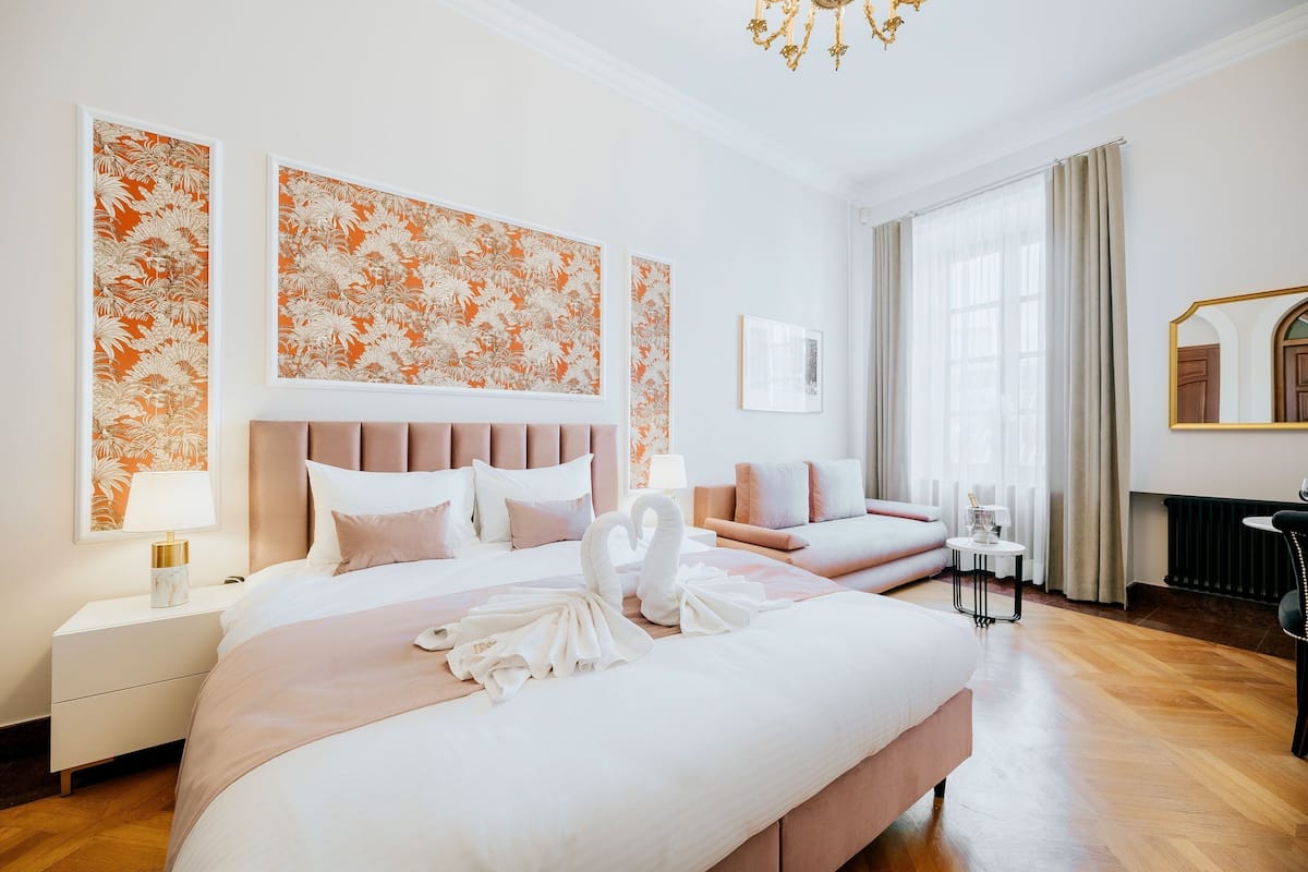 Una habitación de hotel con una cama grande, un sofá más pequeño, una mesita de noche con lámpara y un espejo.