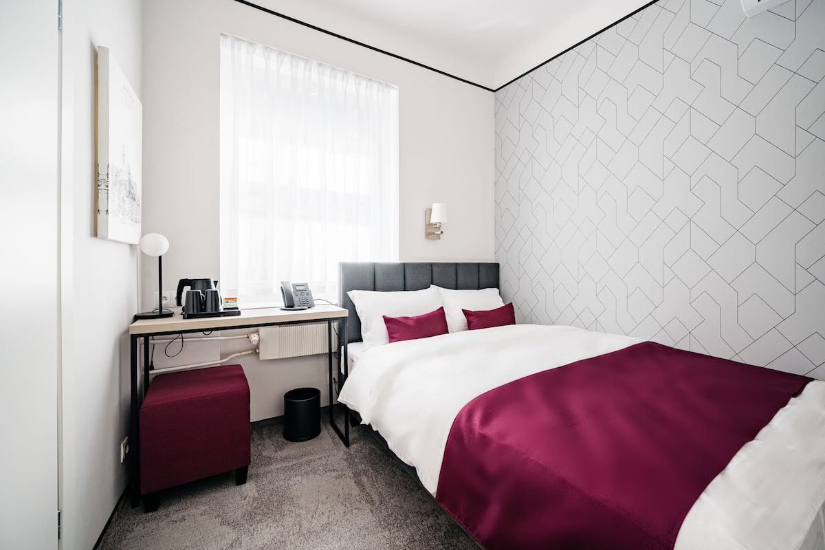 Habitación de hotel con cama, escritorio con lámpara, una silla, un cesto de basura y ventana con cortinas.