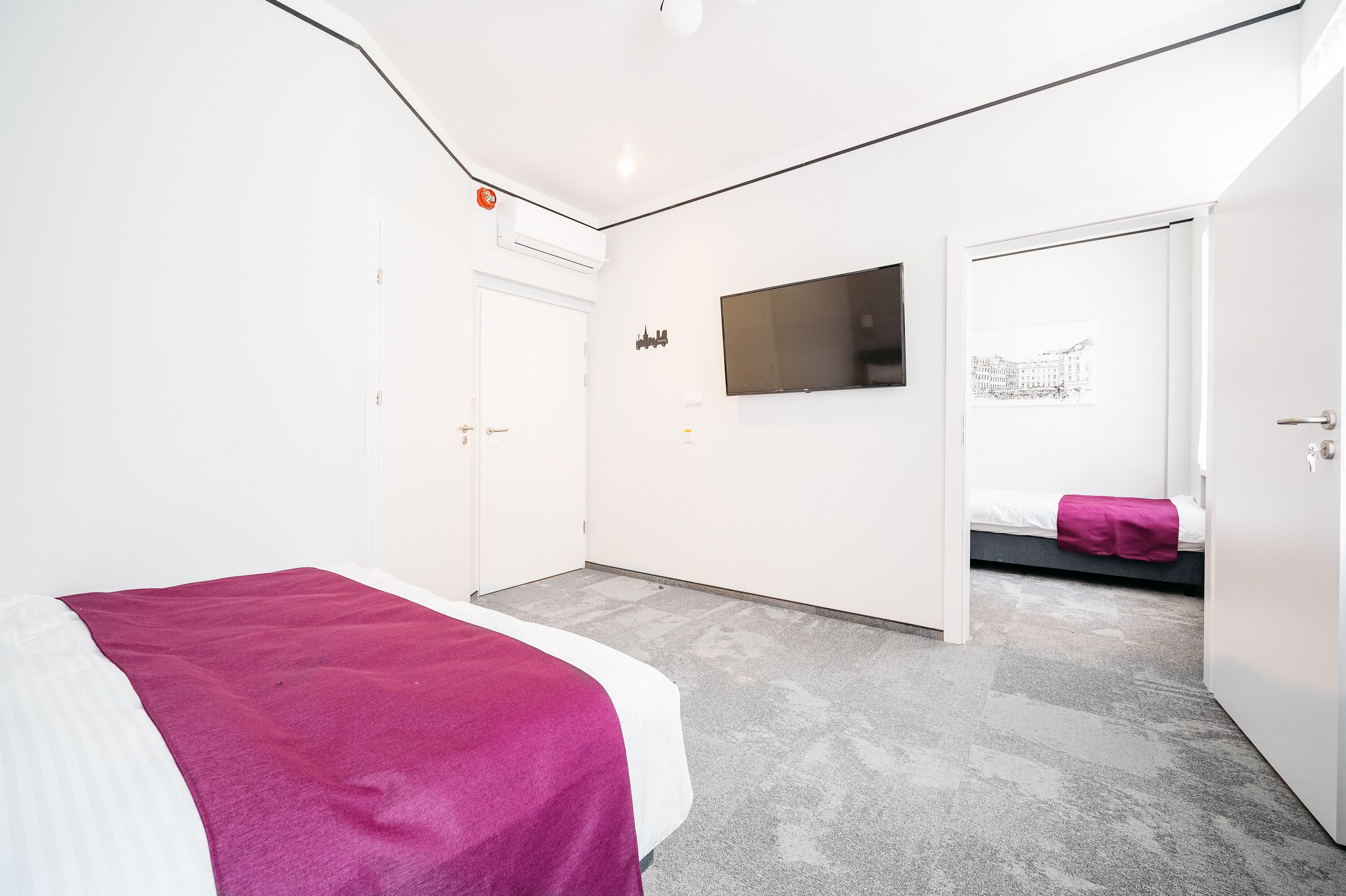 A modern hotel room with a bed, a flat-screen TV, a door leading to another room, and a wall-mounted artwork.