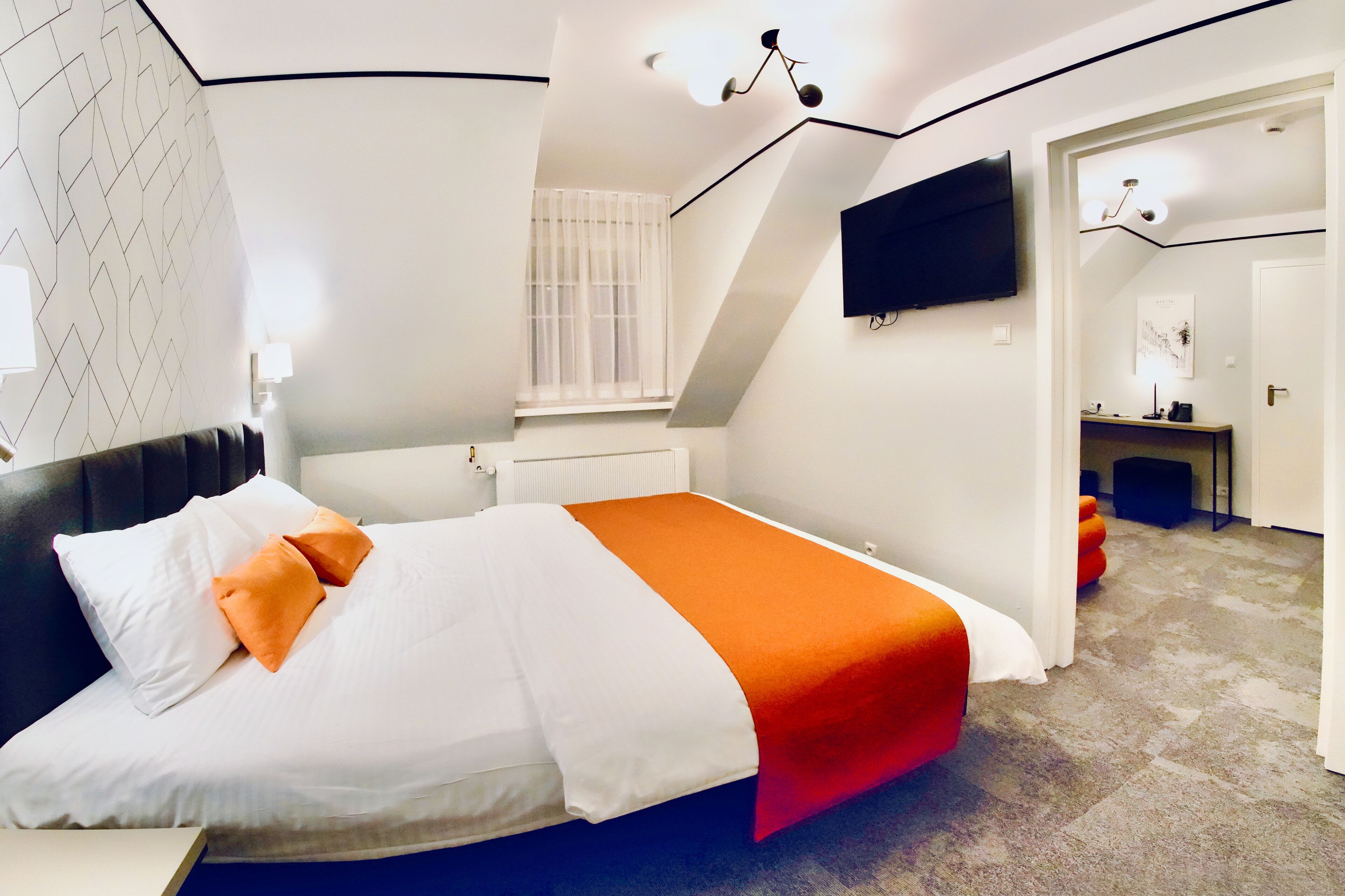 A modern hotel room with a neatly made bed, a flat-screen TV mounted on the wall, and a small desk area with a chair.