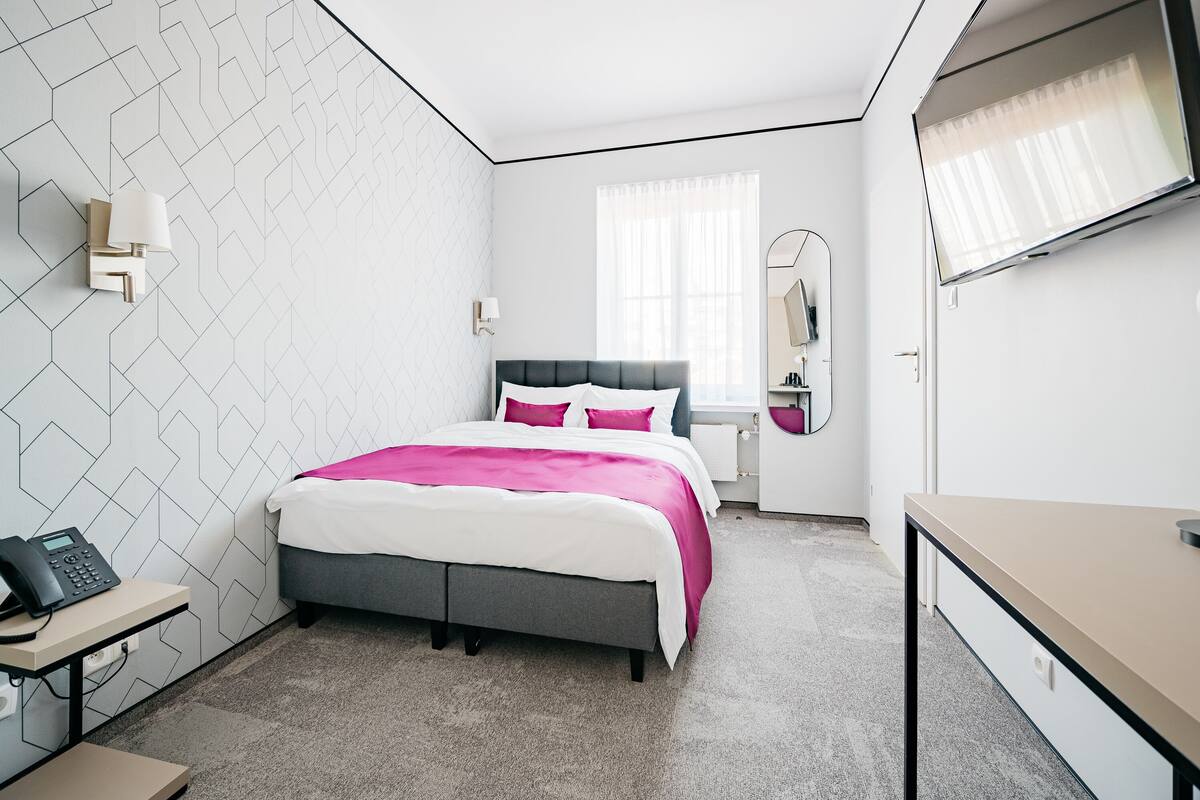 Una habitación de hotel moderna con una pared de diseño geométrico, una cama con ropa de cama blanca y rosa, un escritorio con teléfono y una ventana grande con cortinas translúcidas.