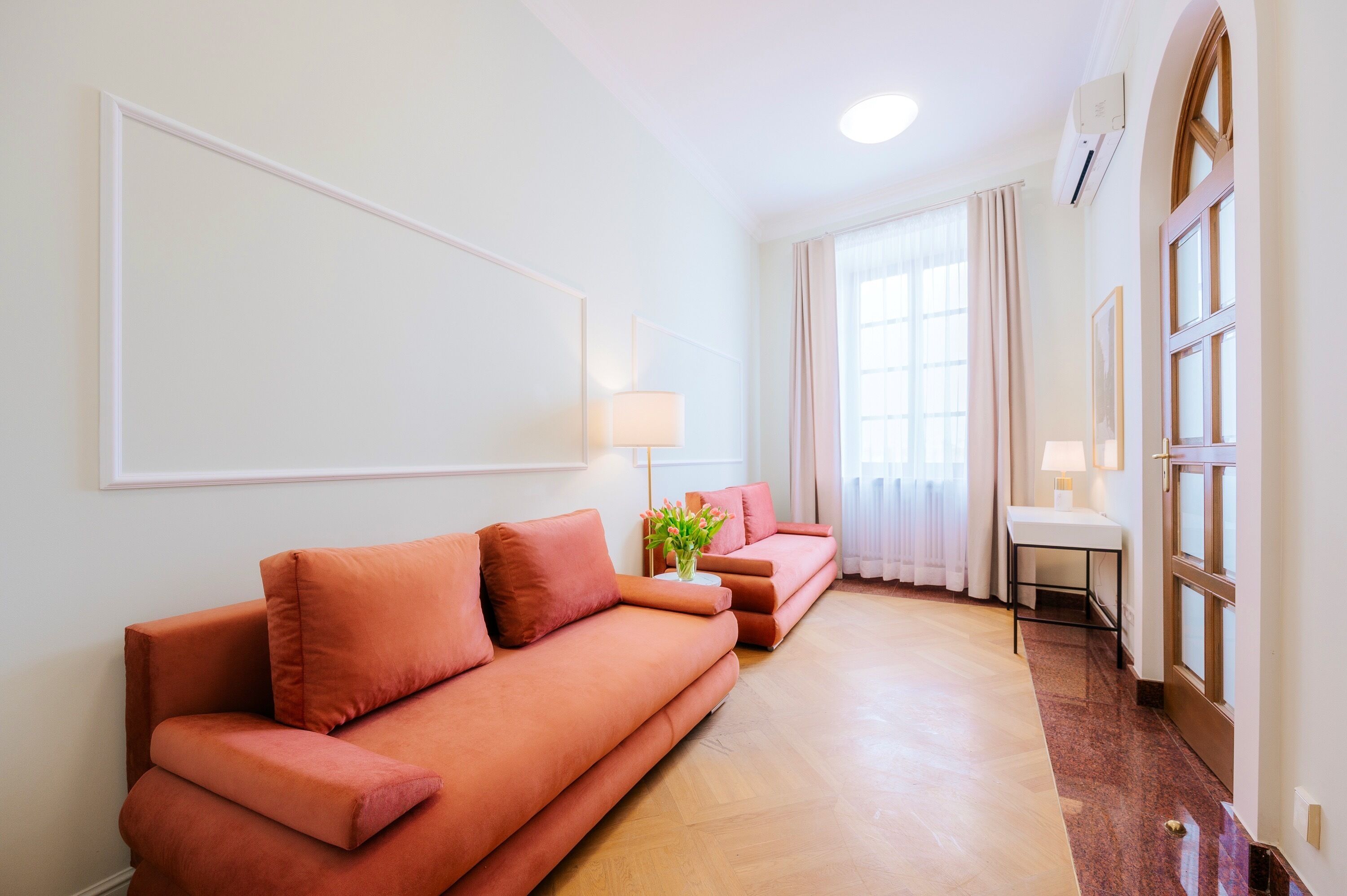Ein modernes Wohnzimmer mit einer Sofagarnitur, einem kleinen Tisch mit Lampe und einem Fenster mit Vorhängen.