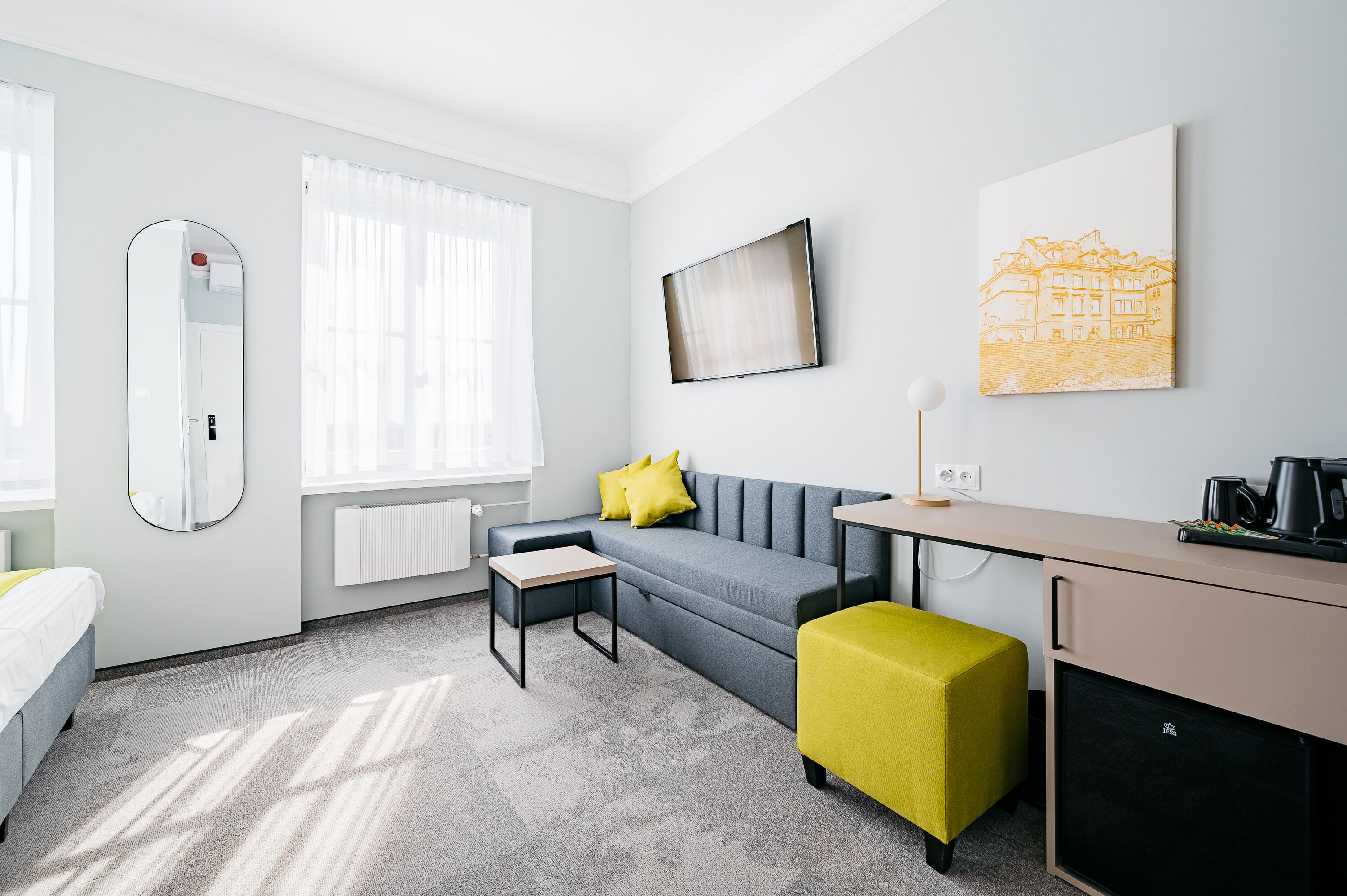 Ett modernt hotellrum med en grå soffa, en gulottoman, ett skrivbord med en kaffebryggare, en stor spegel och ett konstverk som föreställer en stadsbild på väggen.
