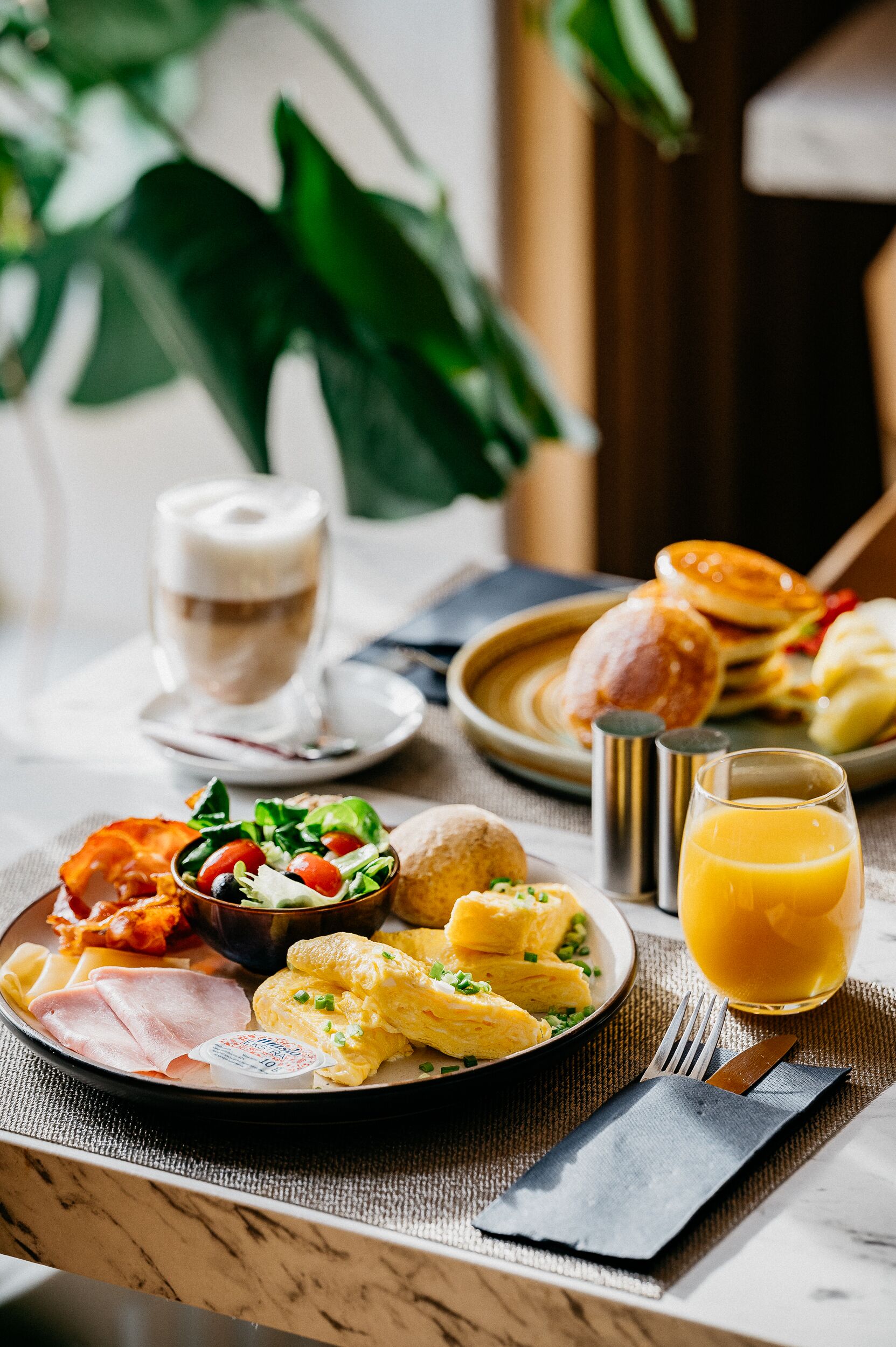 Une table dressée pour le petit-déjeuner avec une assiette d’œufs, de bacon, de jambon et une salade, un verre de jus d’orange et une tasse de café.