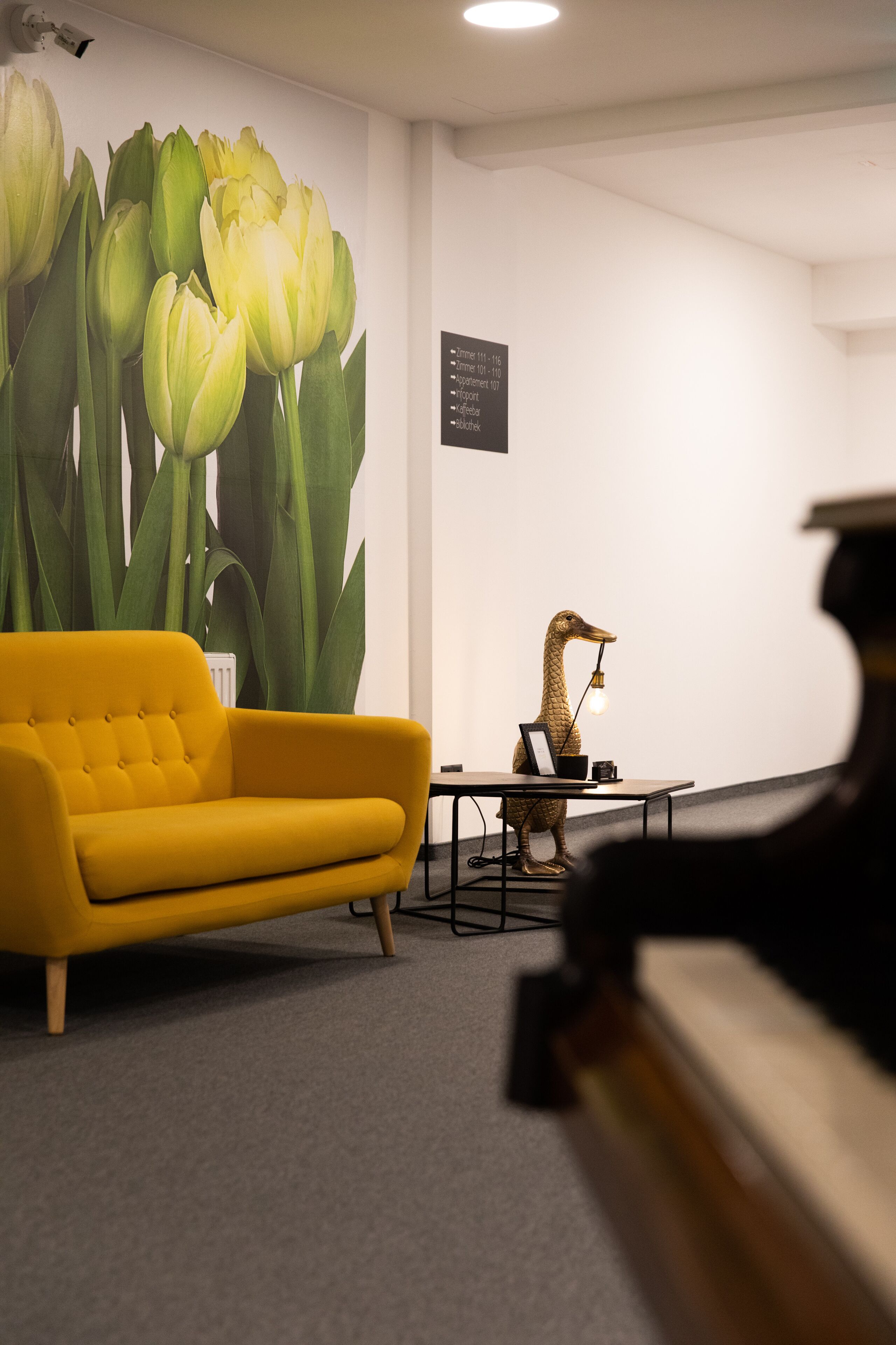 Una habitación con un sofá amarillo, un gran mural de tulipanes, una escultura de un pájaro y un piano.