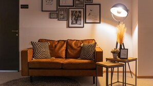 A modern living room with a brown sofa, two patterned cushions, a small wooden side table with a lamp, and a wall adorned with framed pictures. - Self-Check-in Hotel VinoQ Zistersdorf (Zistersdorf)