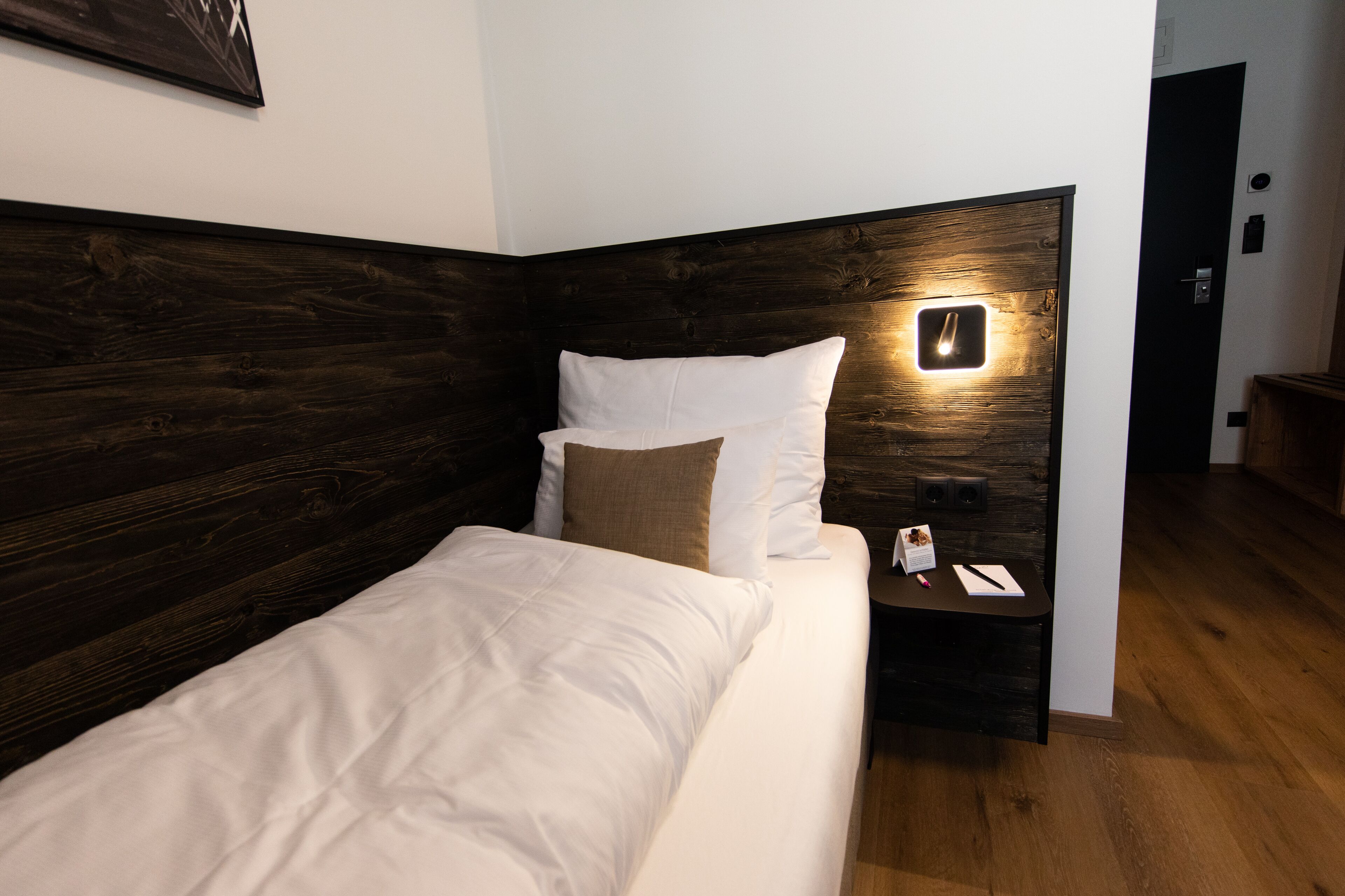 Un lit bien fait, avec une tête de lit en bois, une table de chevet sur laquelle se trouvent un téléphone et un livre, et une lampe fixée au mur.