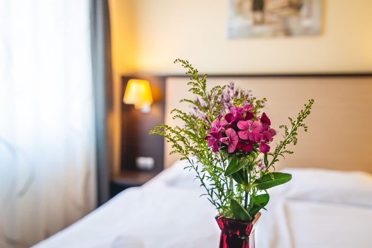 Een bed met witte lakens en een rode vaas met bloemen.