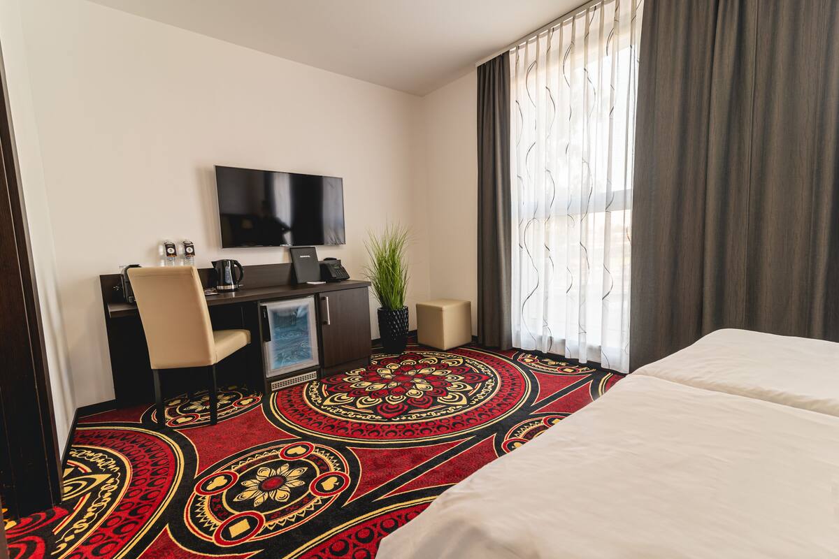Een hotelkamer met een bed, een bureau met een televisie, een plant en een kamerbreed tapijt met dessin.