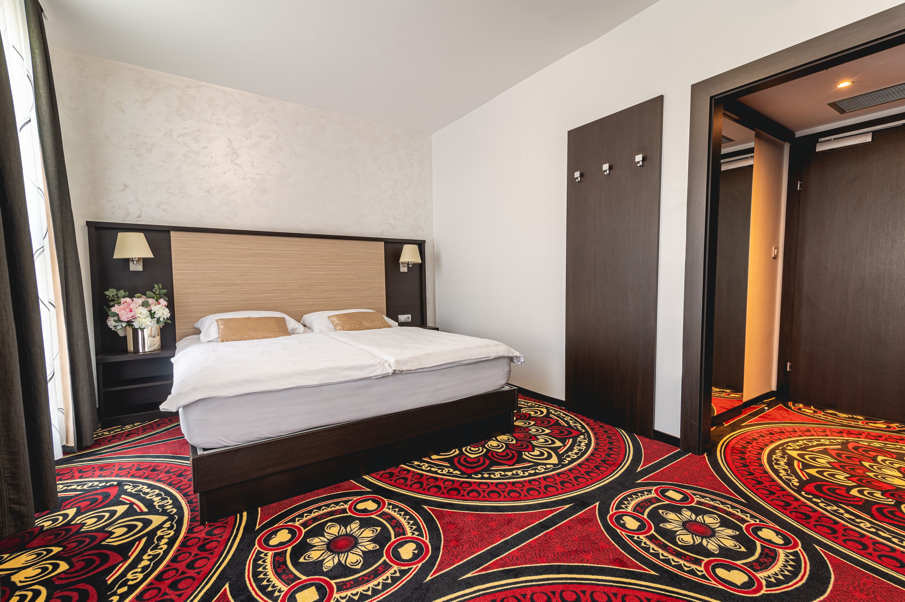 A hotel room with a bed, a nightstand with a lamp, and a door leading to another room.
