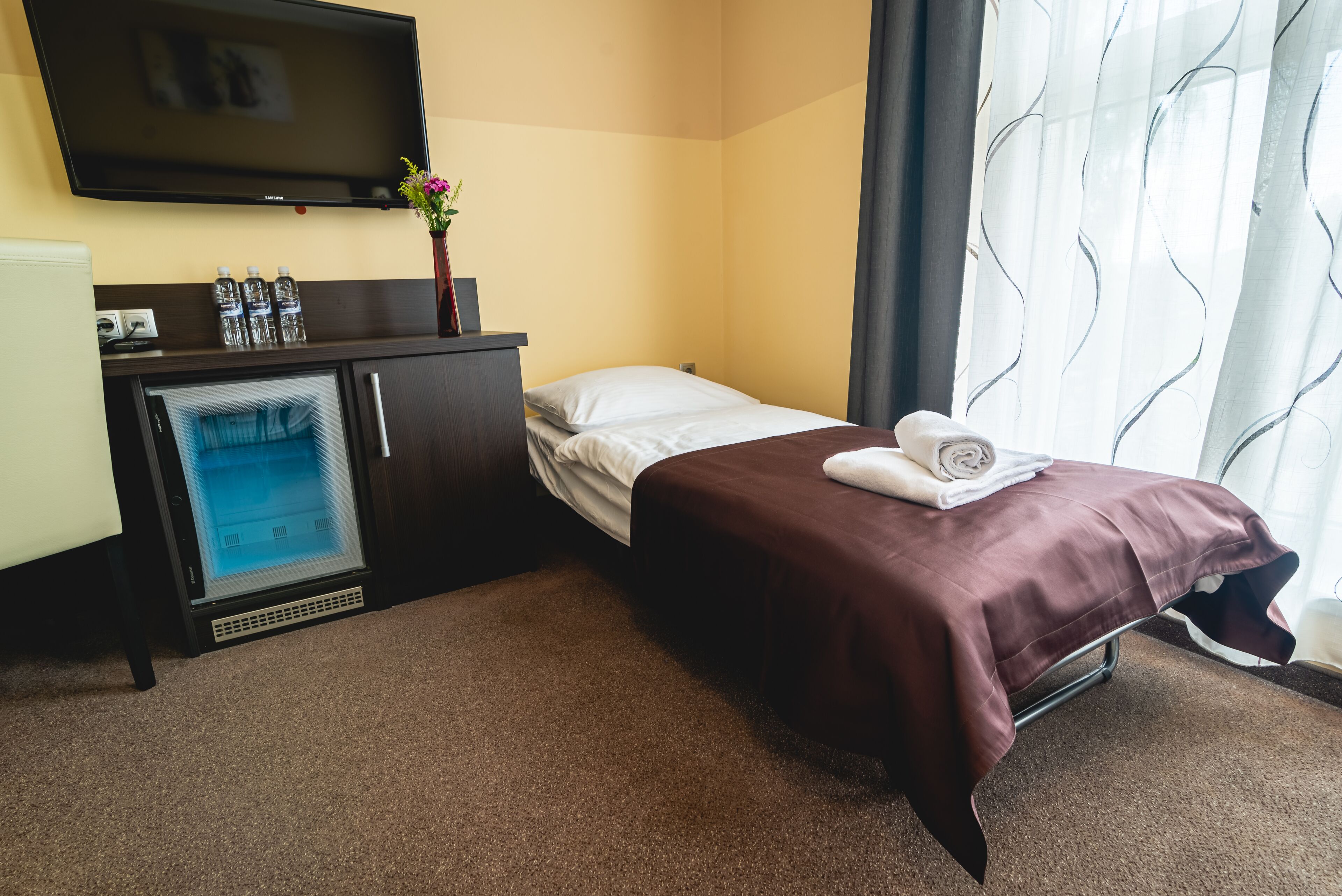 A hotel room with a bed, a TV mounted on the wall, a minibar, and a window with curtains.