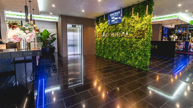 Eine moderne Hotelhalle mit einer Rezeption, einer digitalen Anzeige, einer grünen Wand mit Pflanzen und einem polierten schwarzen Boden.