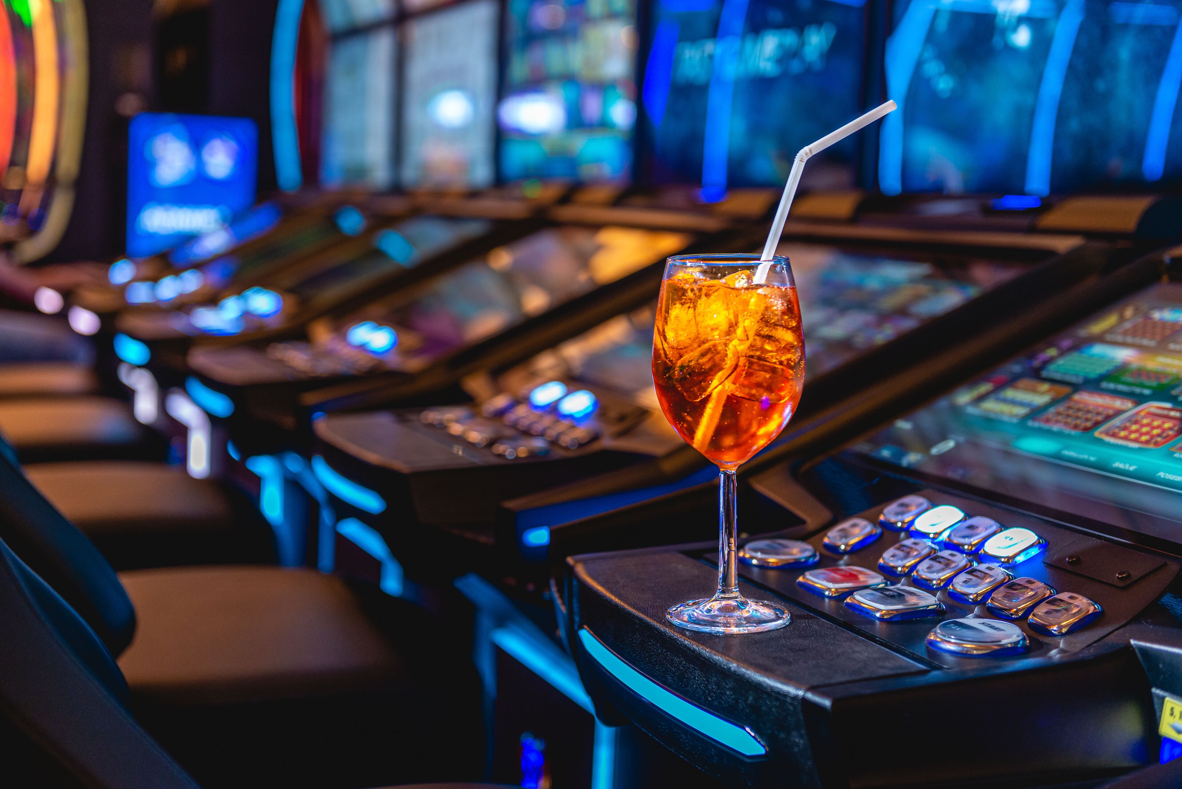 Zona de bar con un vaso de bebida color ámbar con pajita, máquinas tragamonedas iluminadas y una fila de sillas.