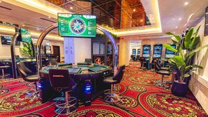 Ein Kasino mit mehreren Spieltischen, einem großen digitalen Roulette-Rad und einem Teppichboden mit aufwendigen Mustern.