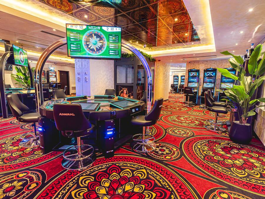Un casino con múltiples mesas de juego, una ruleta digital grande y un suelo alfombrado con diseños elaborados.