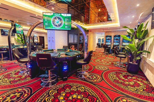 A casino with multiple gaming tables, a large digital roulette wheel, and a carpeted floor with intricate patterns. - Casino & Hotel ADMIRAL Ptuj (Ptuj)