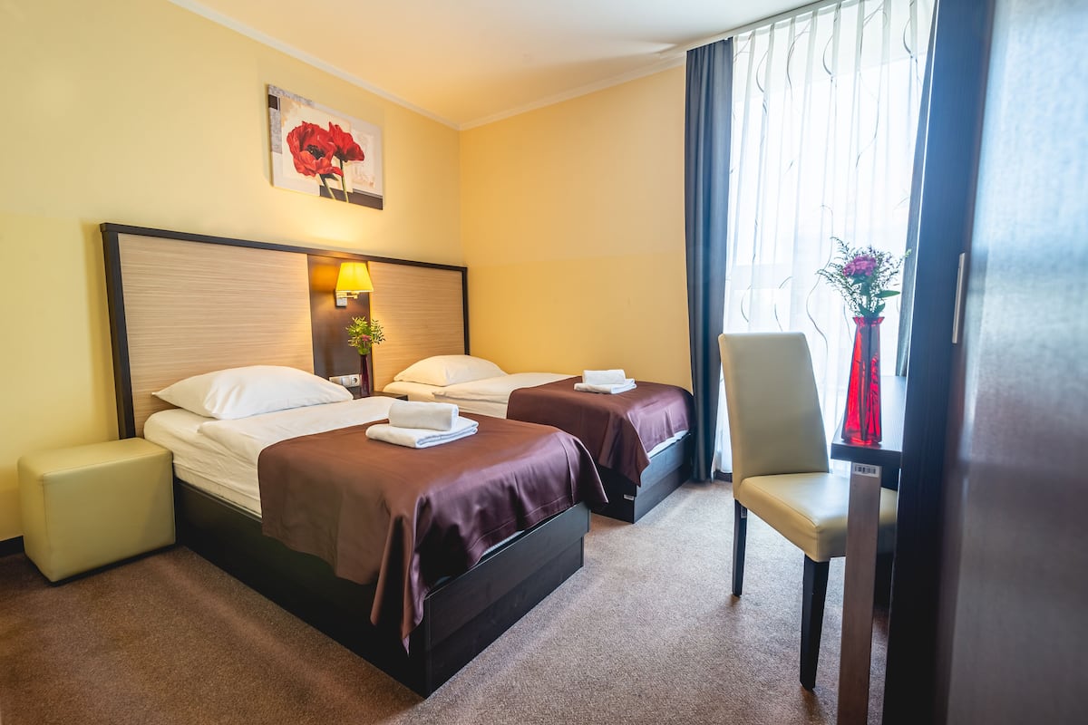 Een hotelkamer met twee bedden, een nachtkastje, een stoel, een lamp, een schilderij en een raam met gordijnen.