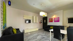 A modern living room with a black sofa, a white dining table, and a TV mounted on the wall.