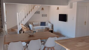 Un coin repas avec une table en bois et des chaises blanches, un escalier menant à un salon équipé d’un canapé et d’un téléviseur, et une porte sur la droite.