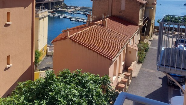 Une ville côtière aux bâtiments aux toits de tuiles rouges, dotée d’un port de plaisance et surplombée d’un ciel bleu azur.
