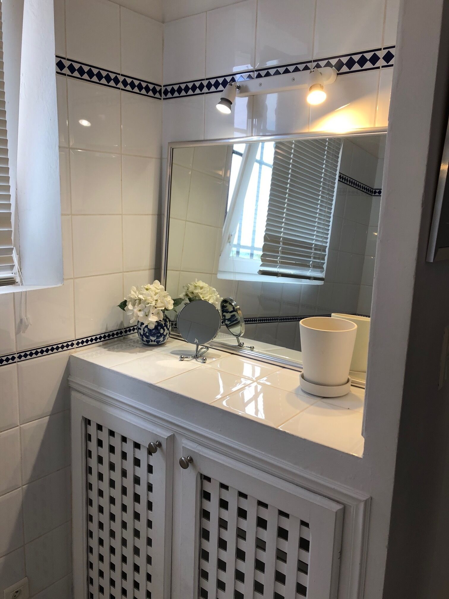 A bathroom with white tiles, a mirror, a sink, and a window with blinds.