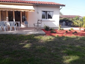 A house with a tiled roof, a covered porch with white plastic chairs, and a small garden area with red mulch and green plants. - Single-storey house 110m², residential area, 500m from town center (Mirande)