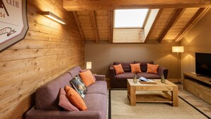 Ein gemütliches Loft-Zimmer mit Holzbalken, einem Oberlicht, zwei Sofas mit Kissen, einem Couchtisch und einem Fernseher auf einem Holzständer.