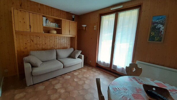 A room with a wooden wall, a sofa, a table with a patterned tablecloth, and a window with curtains. - East-facing studio cabin for 4 guests, pet-friendly (Valloire)
