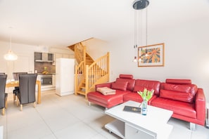 Una sala moderna con un sofá de cuero rojo, una mesa de centro blanca y un comedor con una mesa y sillas de madera. Una escalera conduce a un nivel superior.