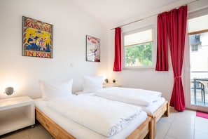 Habitación de hotel con dos camas, una mesita de noche, una ventana con cortinas rojas y dos carteles en la pared.