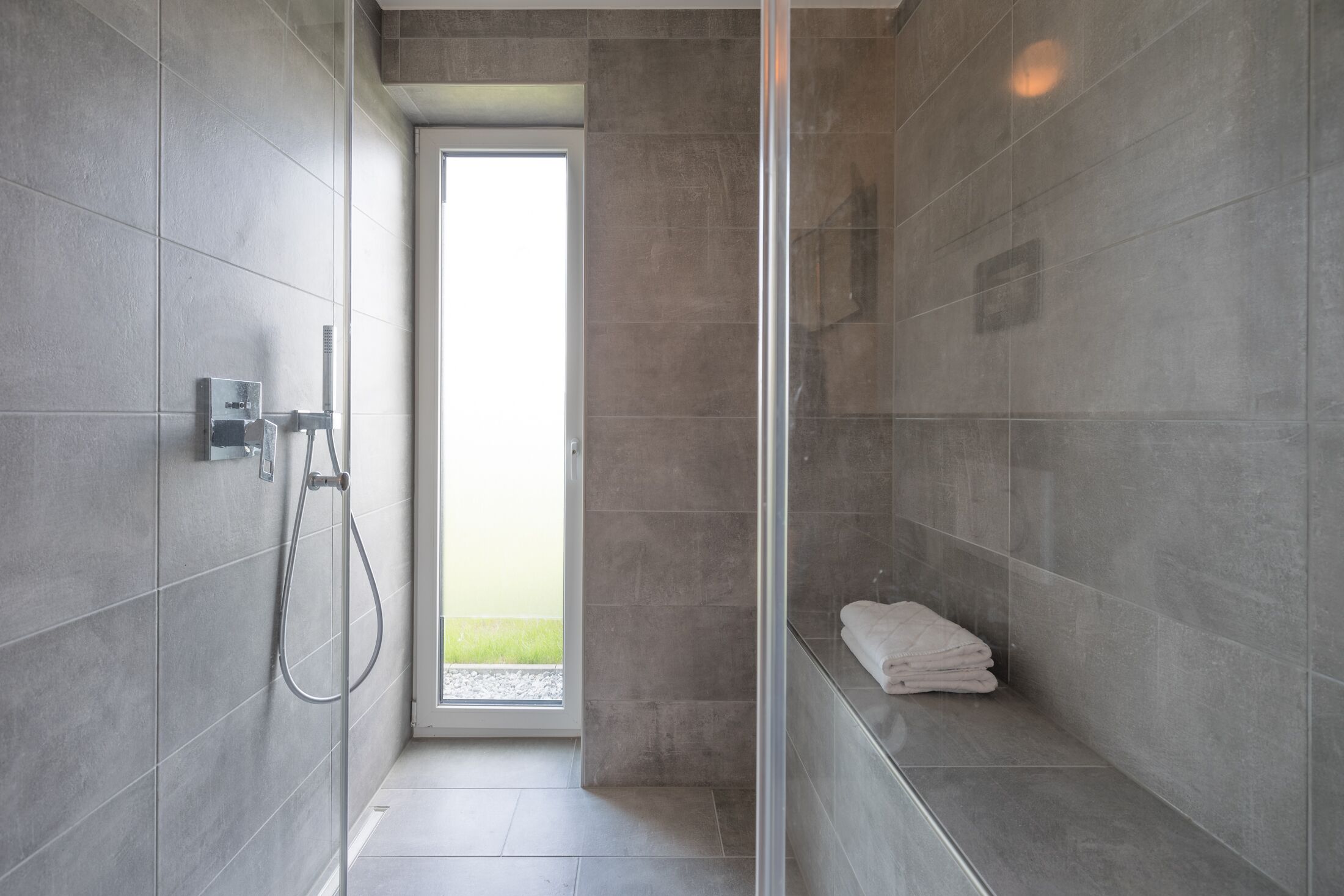 Un espacio de ducha moderno con puerta de vidrio, azulejos grises y una toalla en el suelo.