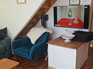 A room with a blue chair, a white table with a bowl, a television displaying a person, and a wooden staircase.