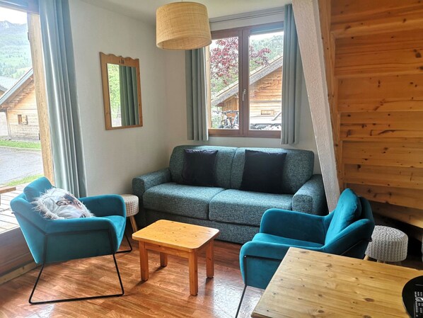 Un salon chaleureux avec un canapé gris, deux fauteuils bleus, une table basse en bois et une petite table en bois. Une fenêtre offre une vue sur un bâtiment en bois et sur la verdure environnante.