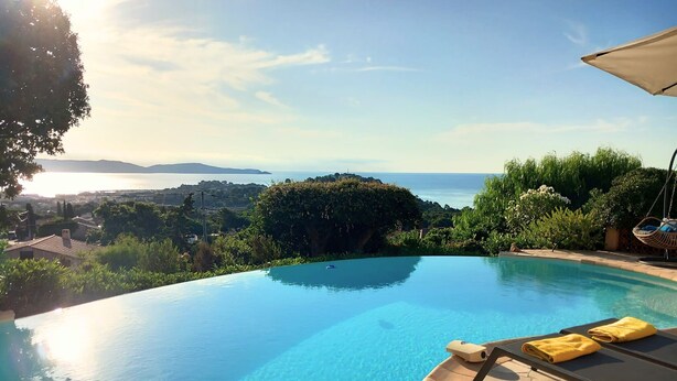 A clear blue infinity pool with a view of the sea and hills, surrounded by lush greenery and a hanging chair. - Villa with heated pool and panoramic views of the Gulf of Saint Tropez (Cavalaire-sur-Mer)