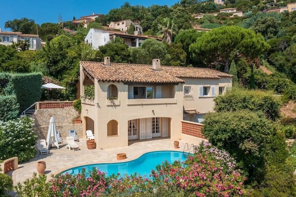 Una casa de dos pisos con piscina, rodeada de abundante vegetación y otras viviendas en una colina.